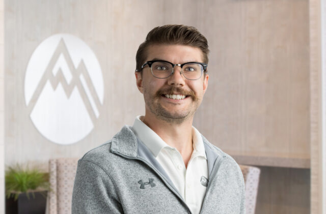 Smiling man with glasses in a light gray jacket, standing in front of a circular logo on a wooden wall.