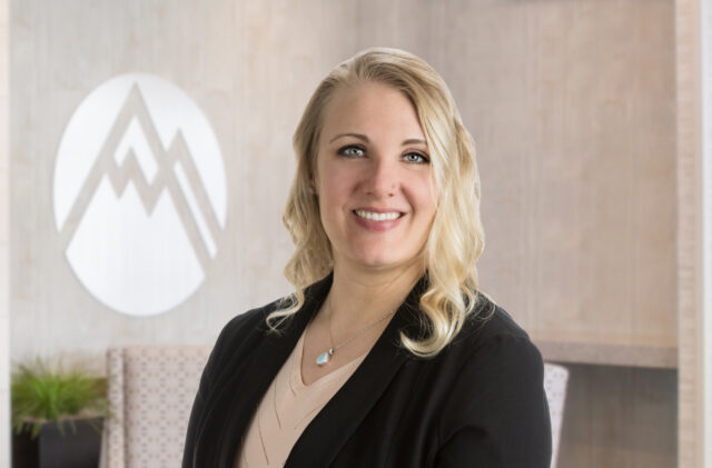 Portrait of a smiling blonde woman in a black blazer and beige top, standing in a modern lobby with a circular logo behind her.
