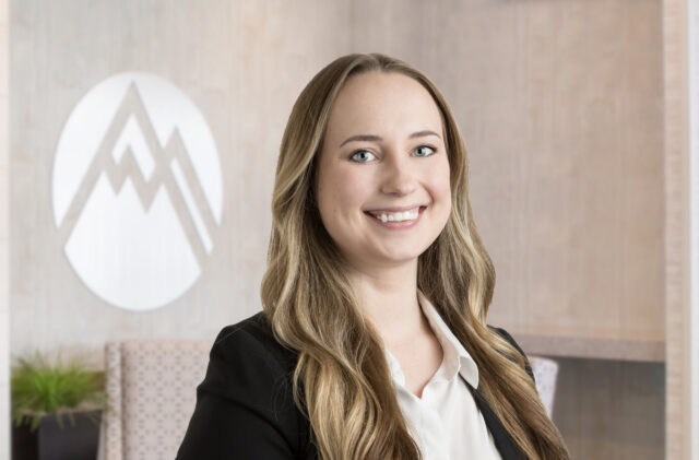Professional woman smiling at camera in office lobby with logo behind her; suited in black blazer and white shirt.