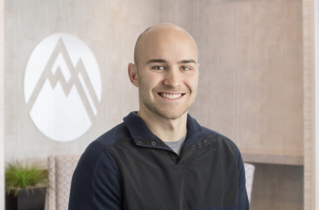 Smiling bald man wearing a dark jacket indoors, with a wooden wall and a large circular logo behind him.