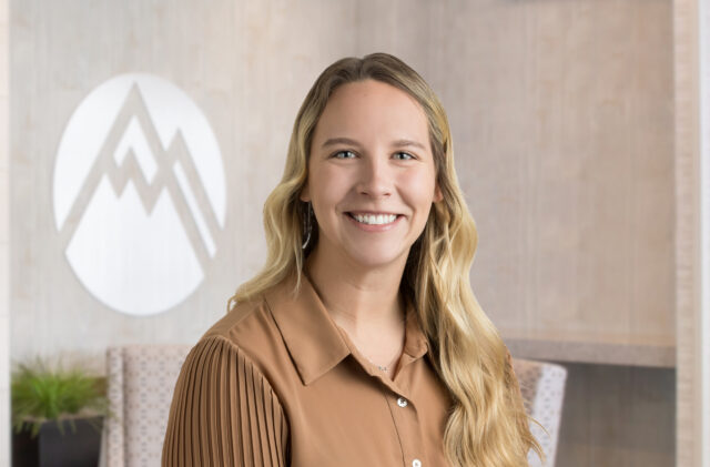 Smiling woman with blond hair in a brown blouse, standing in a modern lobby with a large white logo behind her.