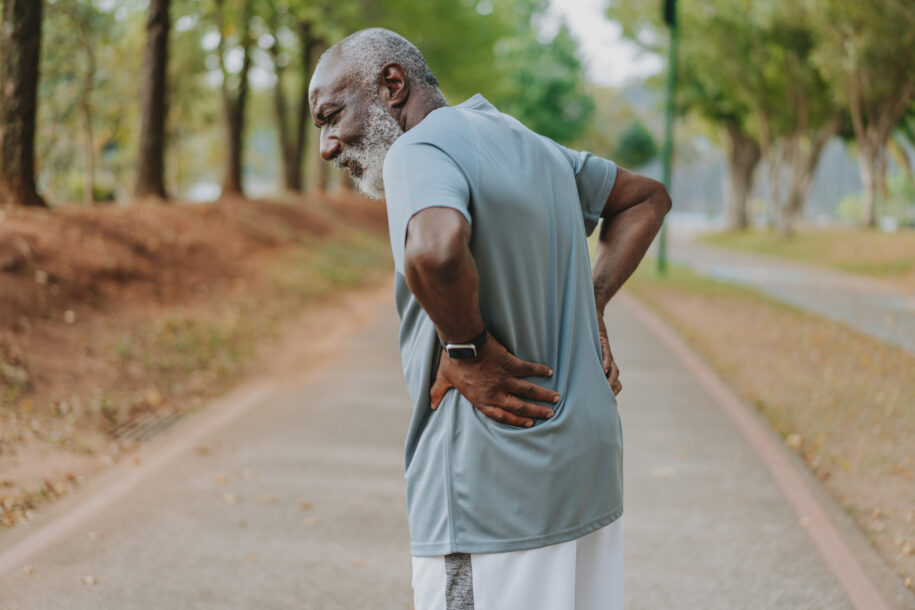 Older man with gray beard in a light blue athletic shirt stands on a park path, hands on lower back, appearing to stretch or ache.