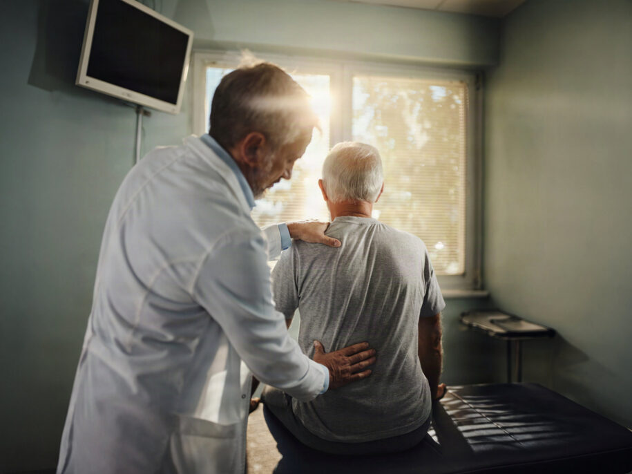 Medical professional in a white coat provides support to an elderly man sitting by a hospital bed as sunlight streams through a window.
